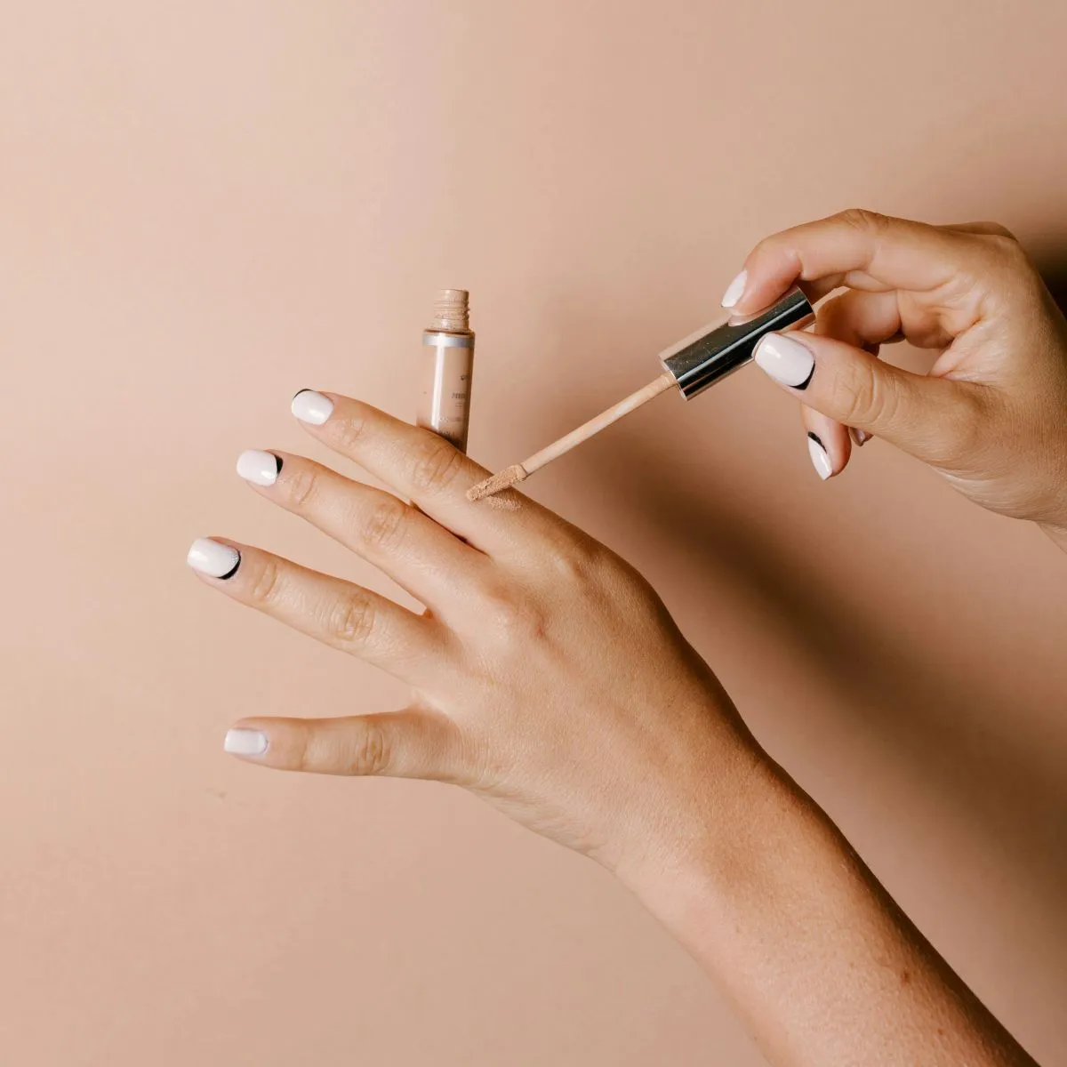 Bej arka planda tırnaklarına beyaz oje üzerine tırnak tasarımı yapılmış, bir elinde kapatıcı diğer elinde kapatıcı fırçasını tutan bir kadının bakımlı elleri